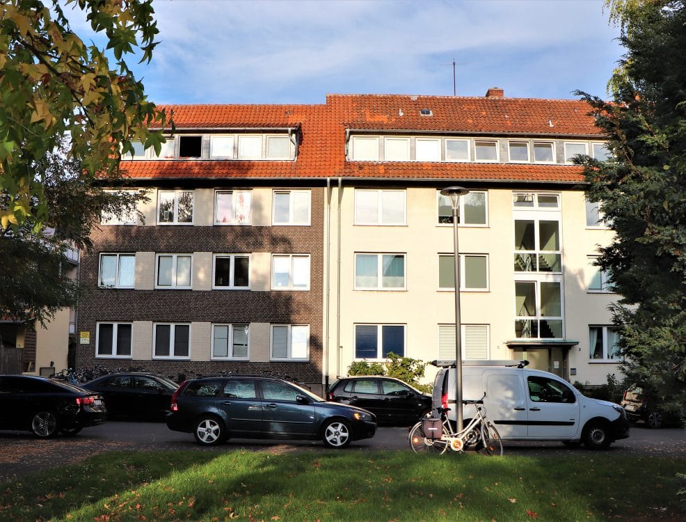 Mehrfamilienhaus mit rotem Ziegeldach, links dunkler Klinkerfassade und rechts hellem Putz, davor parkende Autos und eine Rasenfläche.