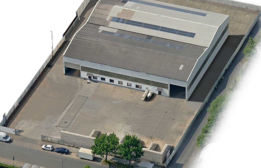 Schräges Luftbild einer Gewerbehalle mit Satteldach und Dachverglasung, umgeben von einem umzäunten, betonierten Hof.