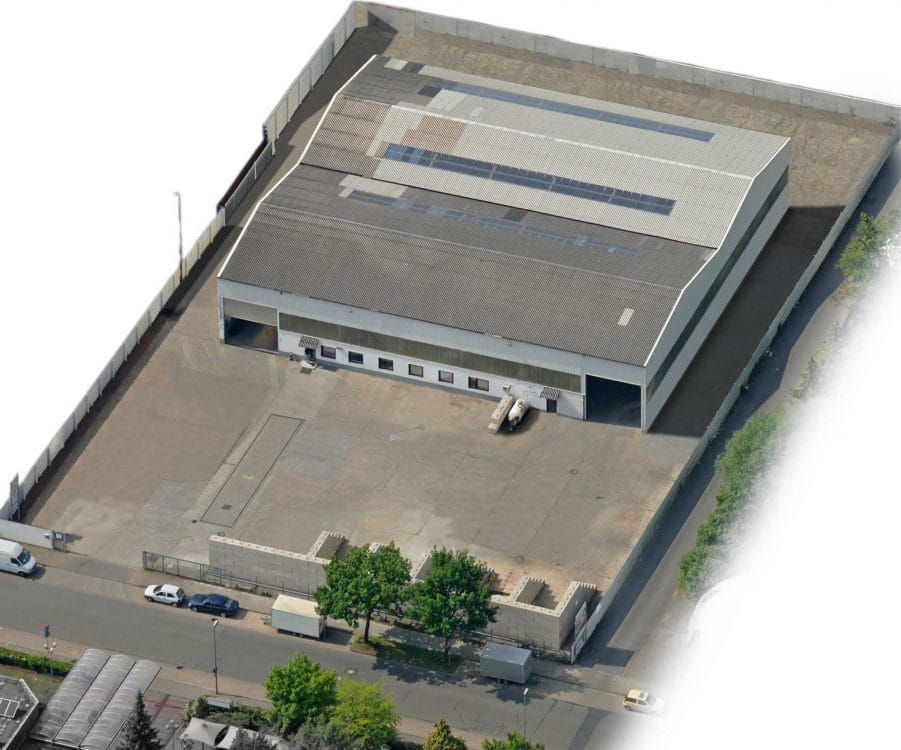 Schräges Luftbild einer Gewerbehalle mit Satteldach und Dachverglasung, umgeben von einem umzäunten, betonierten Hof.