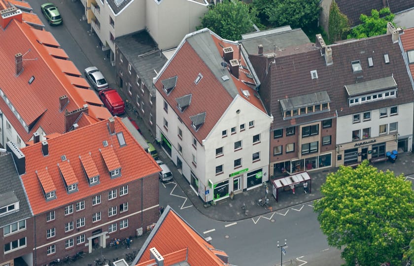 Schräges Luftbild einer Innenstadtbebauung mit einem weißen Giebelhaus und Ladenlokal an einer Straßenecke, ringsum rote Ziegeldächer.