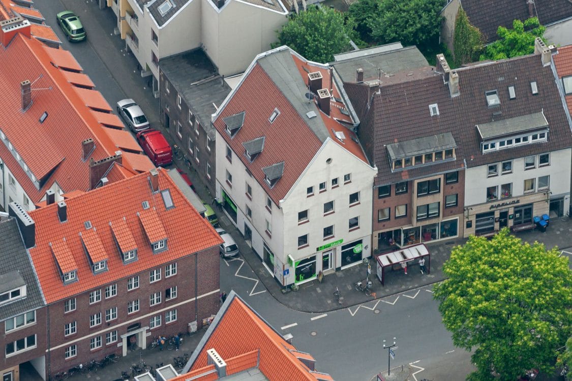 Schräges Luftbild einer Innenstadtbebauung mit einem weißen Giebelhaus und Ladenlokal an einer Straßenecke, ringsum rote Ziegeldächer.