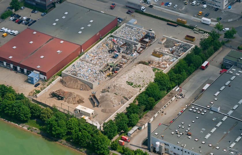 Luftbild eines Gewerbeareals mit Lagerboxen für Schüttgut, Materialhalden und angrenzenden Hallen, links ein Gewässer.