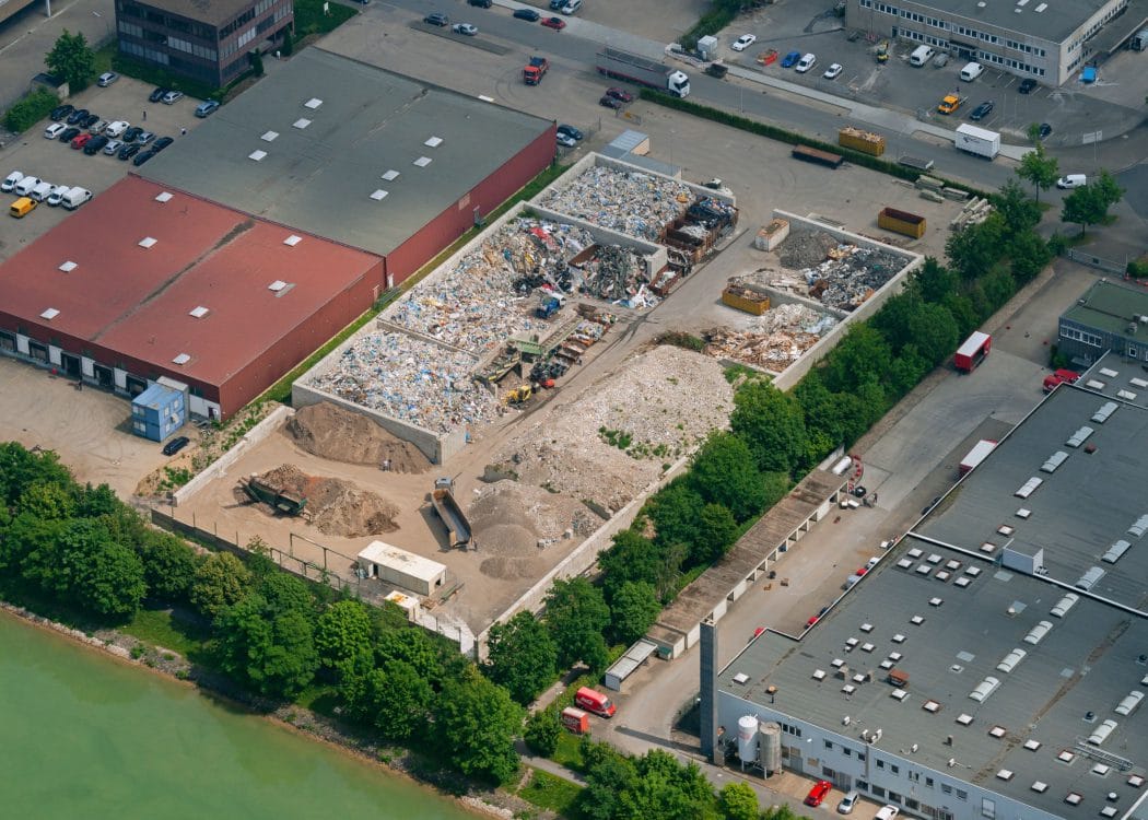Luftbild eines Gewerbeareals mit Lagerboxen für Schüttgut, Materialhalden und angrenzenden Hallen, links ein Gewässer.