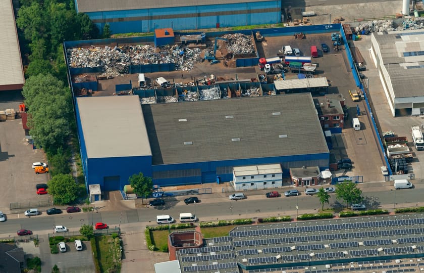 Luftbild einer blau gefassten Gewerbehalle mit angrenzendem Hof, Sortierboxen und Lkw-Stellflächen, unten ein Dach mit Photovoltaikmodulen.