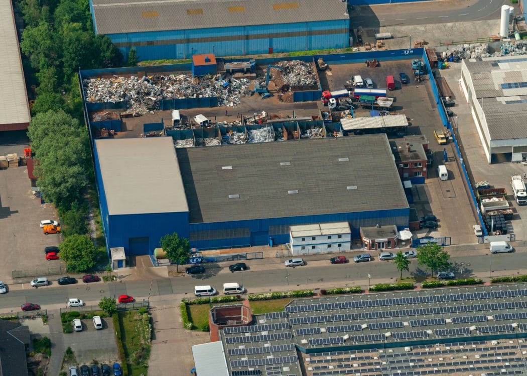 Luftbild einer blau gefassten Gewerbehalle mit angrenzendem Hof, Sortierboxen und Lkw-Stellflächen, unten ein Dach mit Photovoltaikmodulen.
