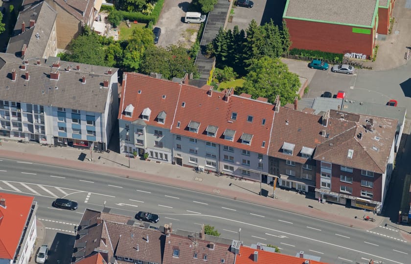 Luftbild einer geschlossenen Häuserzeile mit roten Ziegeldächern und Dachgauben entlang einer breiten, mehrspurigen Straße.