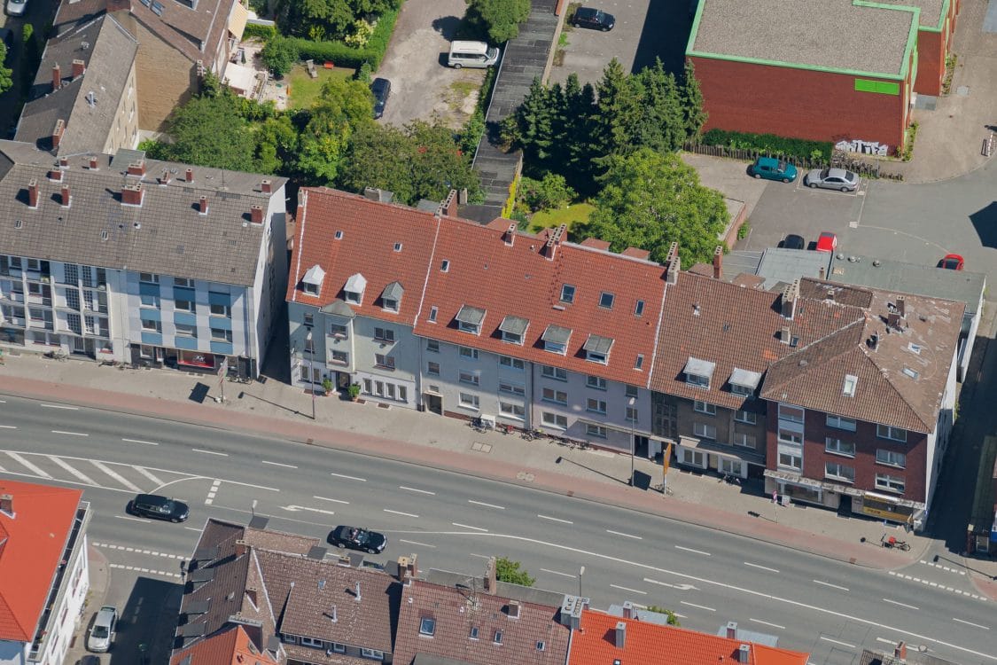 Luftbild einer geschlossenen Häuserzeile mit roten Ziegeldächern und Dachgauben entlang einer breiten, mehrspurigen Straße.