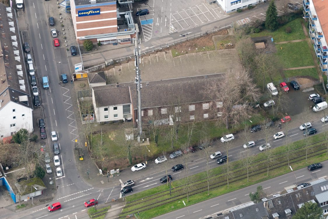 Schräges Luftbild einer langgestreckten Halle mit Satteldach und weißen Toren, daneben ein Wohnhaus und eine Straße mit parkenden Autos.