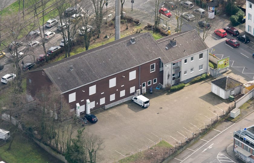 Luftbild einer langgestreckten Halle mit dunklem Satteldach und angebautem weißem Wohnhaus, davor eine weite befestigte Hoffläche.