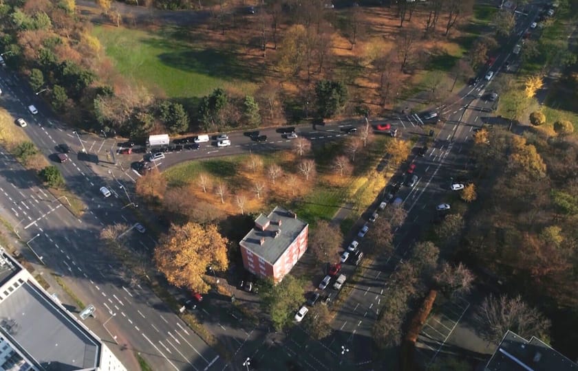 Drohnenaufnahme einer großen Straßenkreuzung mit Grüninseln, in deren Mitte ein freistehendes Mehrfamilienhaus zwischen herbstlichen Bäumen steht.