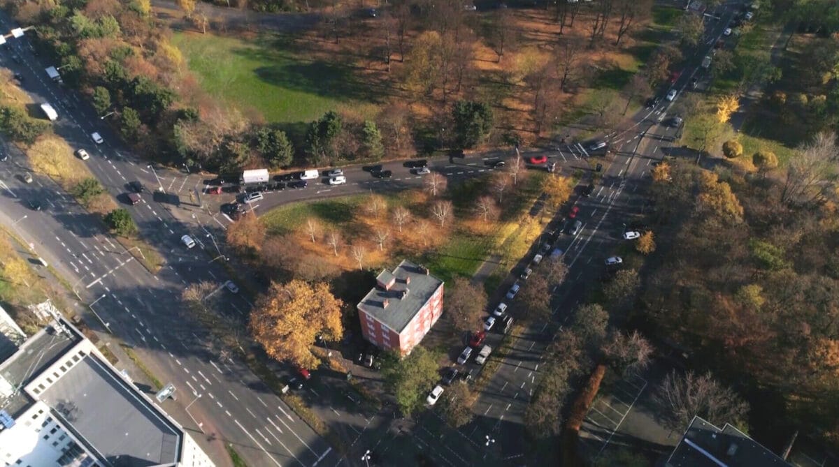 Drohnenaufnahme einer großen Straßenkreuzung mit Grüninseln, in deren Mitte ein freistehendes Mehrfamilienhaus zwischen herbstlichen Bäumen steht.